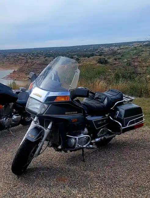 A black Honda Goldwing motorcycle is parked on gravel overlooking a scenic, hilly landscape with a body of water in the…