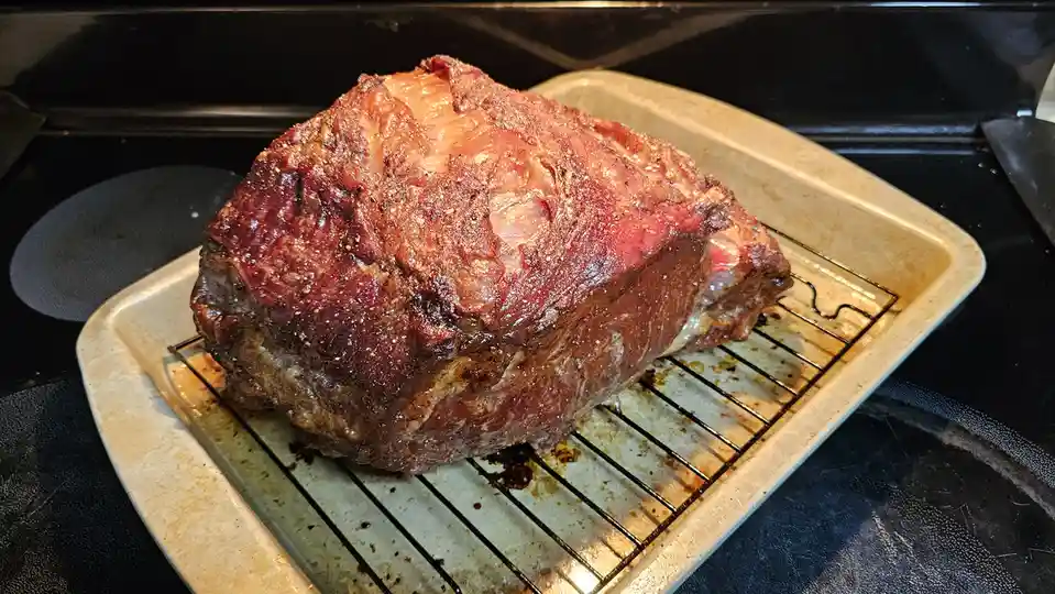 A large, roasted prime rib roast with a browned, seasoned crust sits on a wire rack inside a beige baking pan, which rests…