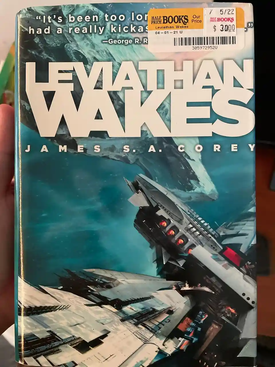 A person holds a copy of James S. A. Corey's book 'Leviathan Wakes', featuring a space battle scene with ships and a large…