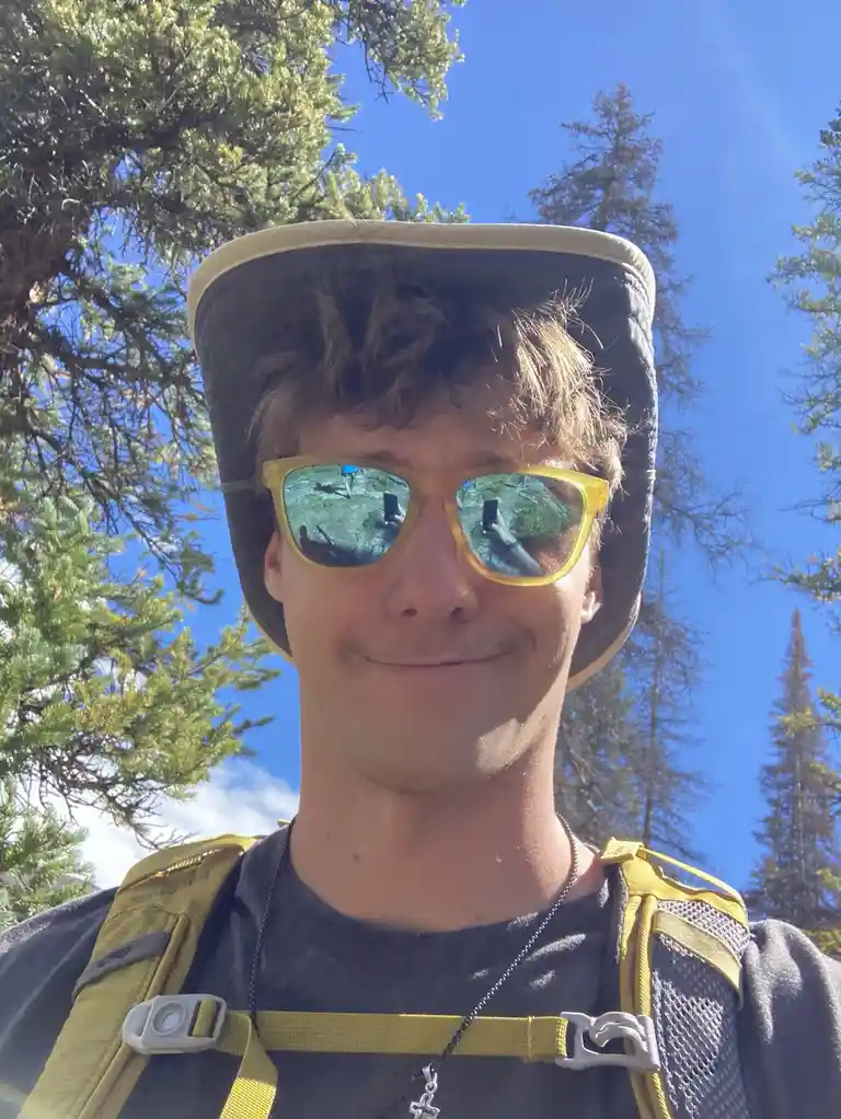 A young man with curly brown hair, wearing a wide-brimmed hat and yellow-framed sunglasses, takes a selfie outdoors. He is…