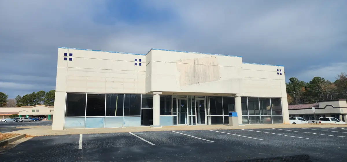 An empty, beige commercial building with large glass windows and doors, a faded sign area, and an empty parking lot under a…