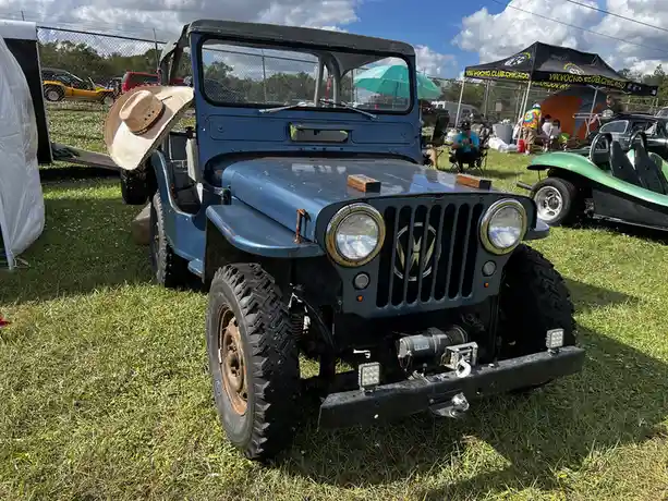 A blue, vintage Jeep with a large sombrero perched on its passenger-side door, parked on grass at an outdoor car show. The…