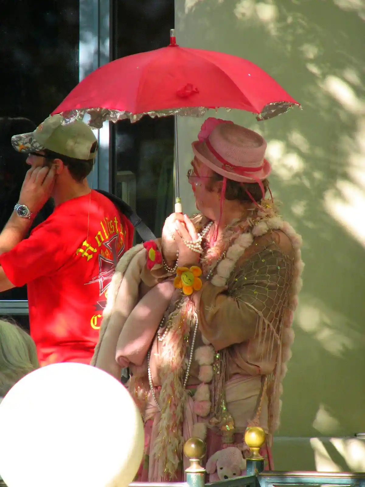 A person in a flamboyant pink outfit with a matching hat and multiple necklaces holds a red umbrella while standing next to…