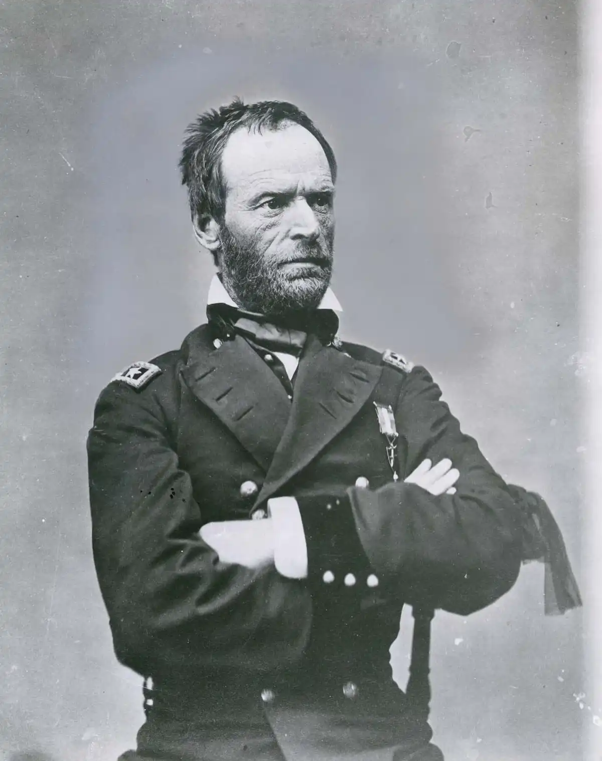 A black-and-white historical portrait of a bearded man in a military uniform with crossed arms, looking sternly off to the…