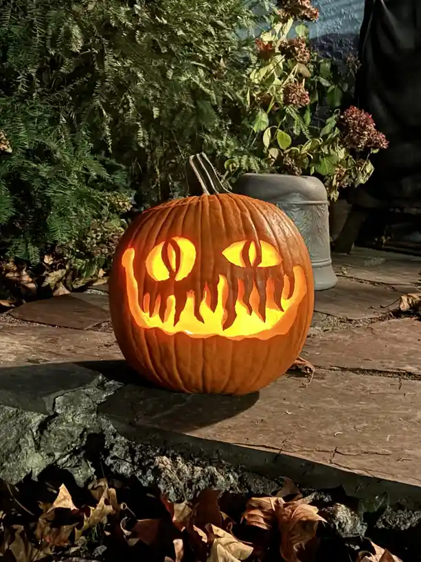 A carved Halloween pumpkin with glowing eyes and a jagged mouth sits on a stone patio, surrounded by greenery and fallen…