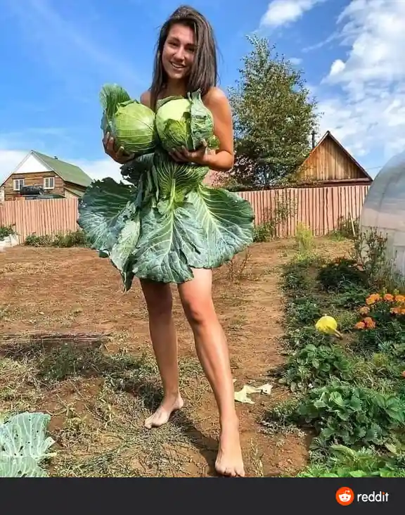 A woman stands barefoot in a garden, smiling while holding two large cabbages, with the lower half of her body covered by a…