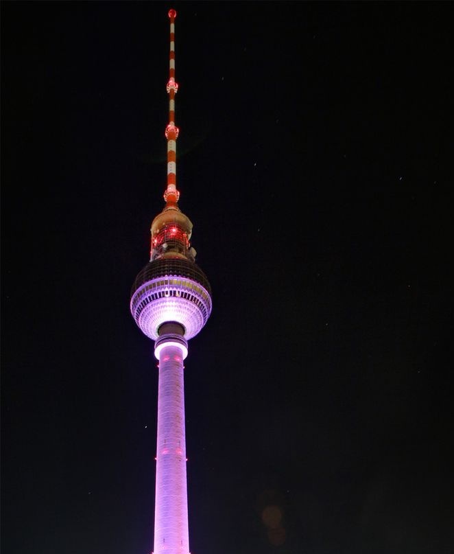 A nighttime photo of the Berlin TV Tower (Fernsehturm) illuminated in purple and red lights against a dark sky. The tower's…