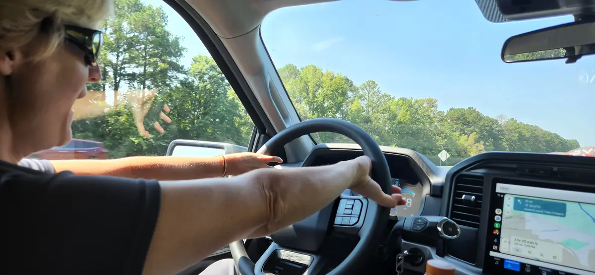 A person with short blonde hair and sunglasses is driving a car, hands on the steering wheel, looking ahead at a road lined…