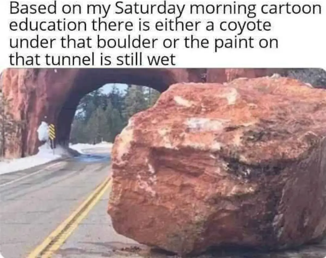 A large reddish-brown boulder blocks a paved road with yellow double lines, directly in front of a tunnel carved into a rock…