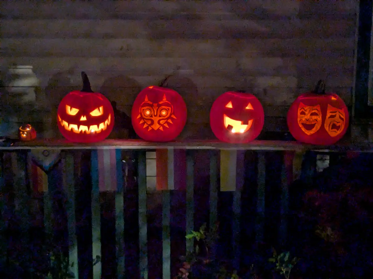 Four glowing jack-o'-lanterns sit in a row on a porch railing at night, each carved with a different face: a classic scary…
