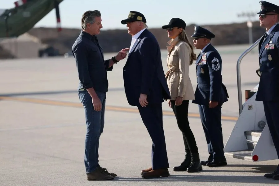 A man in a dark jacket and jeans points at a man in a dark suit and a 'MAKE AMERICA GREAT AGAIN' cap. Behind them, a woman…