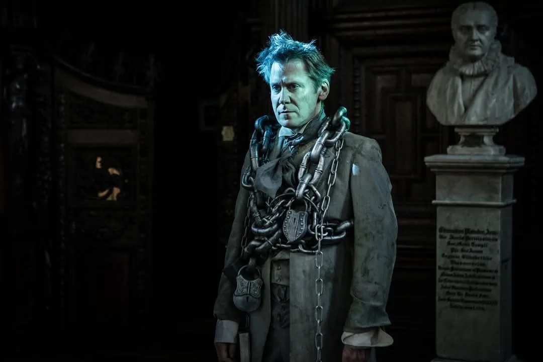 A man in a dark, gothic setting is heavily shackled with chains around his neck, torso, and arms, wearing a tattered coat…