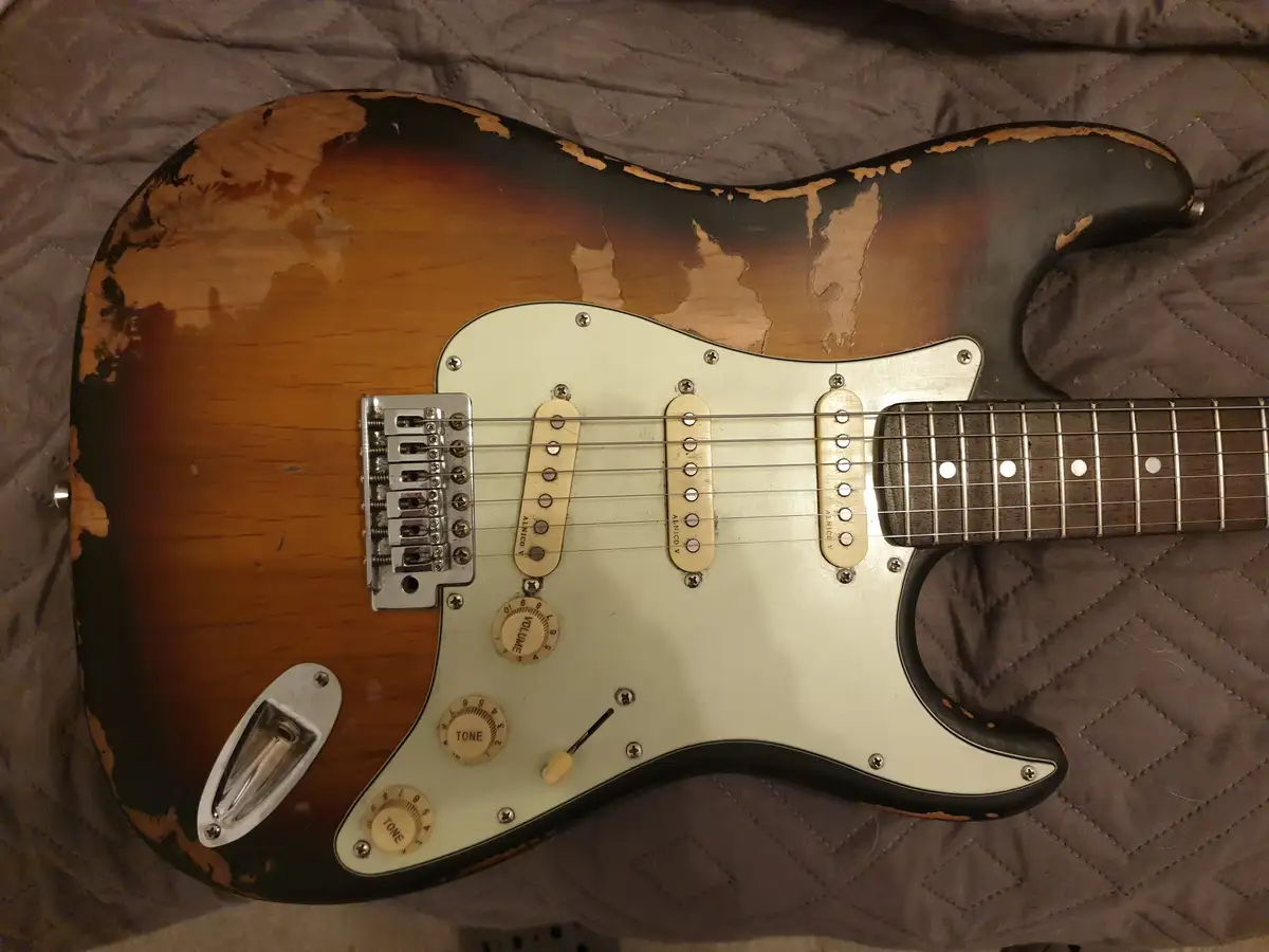 A sunburst-finish electric guitar with heavily worn and peeling paint on its body, revealing the wood underneath, is laid on…