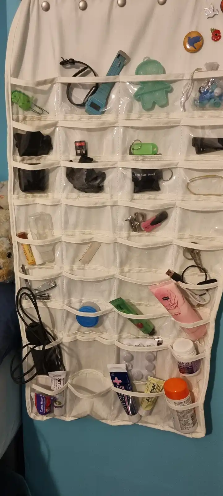 A white over-the-door organizer with clear pockets is filled with various personal items including medicines, a CPR face…