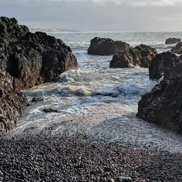 A scenic photo of ocean waves crashing against dark, jagged rocks on a pebbled shore under an overcast sky. The overlaid…