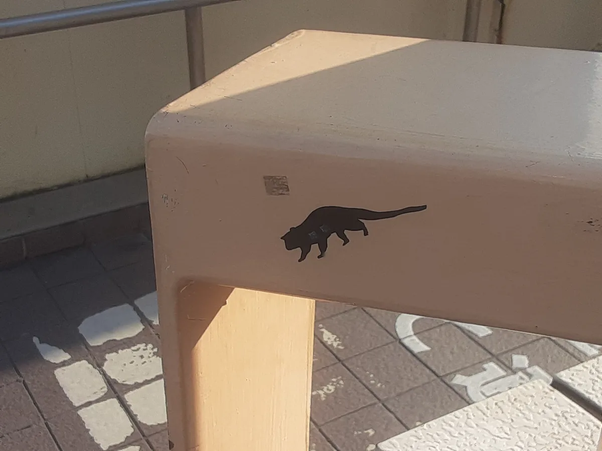 A close-up photo shows a black silhouette decal of a walking cat affixed to the side of a beige outdoor bench. The bench is…