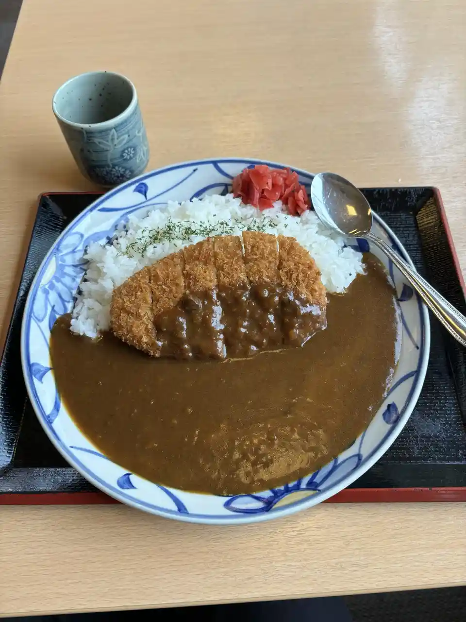 A plate of Japanese curry rice with a breaded cutlet, white rice, pickled ginger, and a spoon, served on a wooden table with…