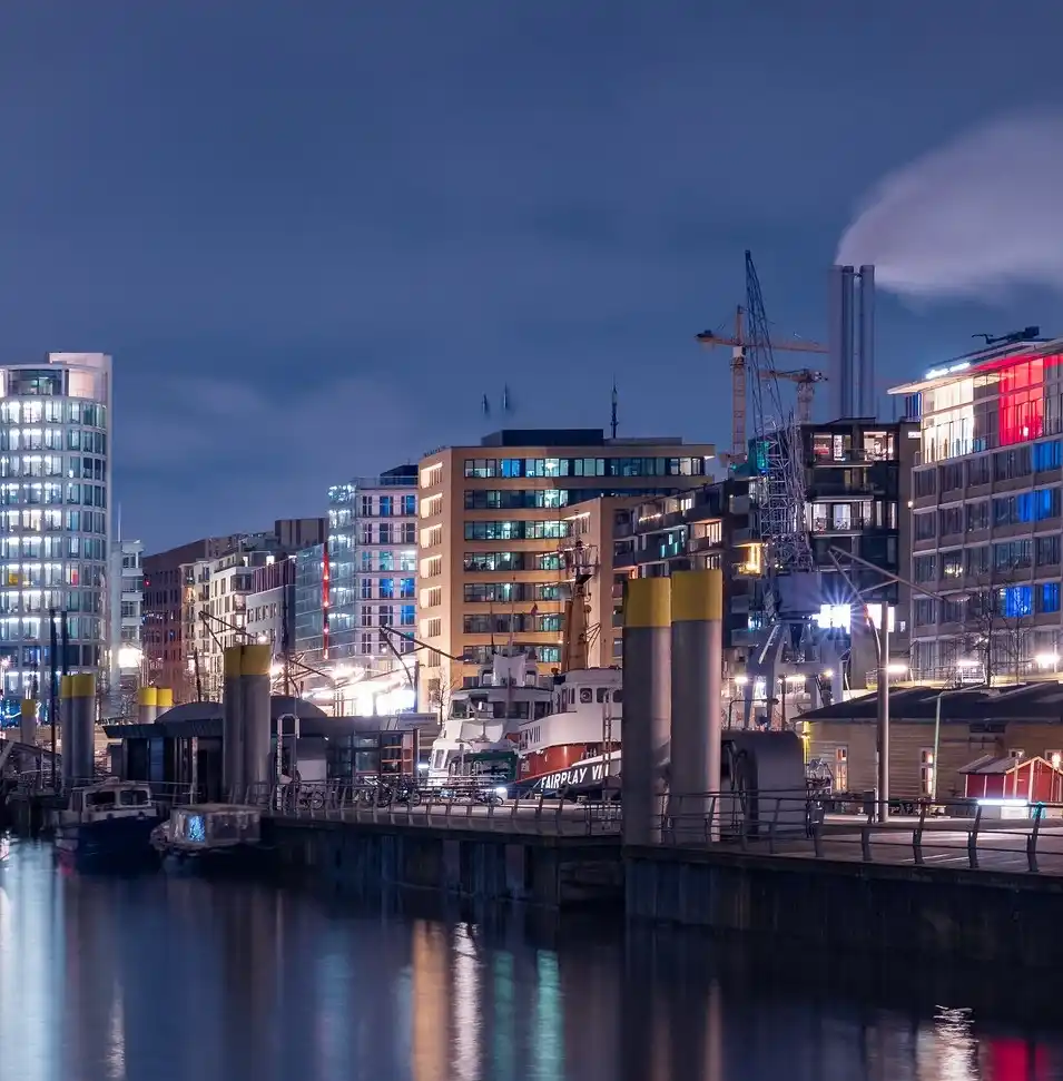 A nighttime city harbor scene with illuminated modern buildings, cranes, and boats on calm water reflecting lights. A white…