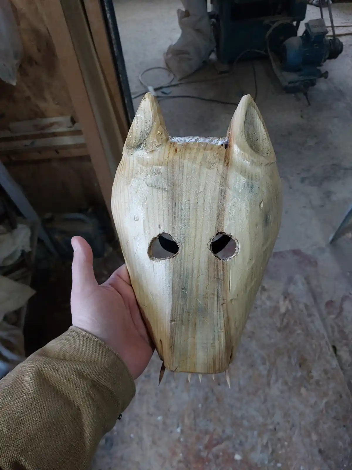 A hand holds up a crudely carved wooden wolf mask with pointed ears, eye holes, and tooth-like protrusions, in a workshop…