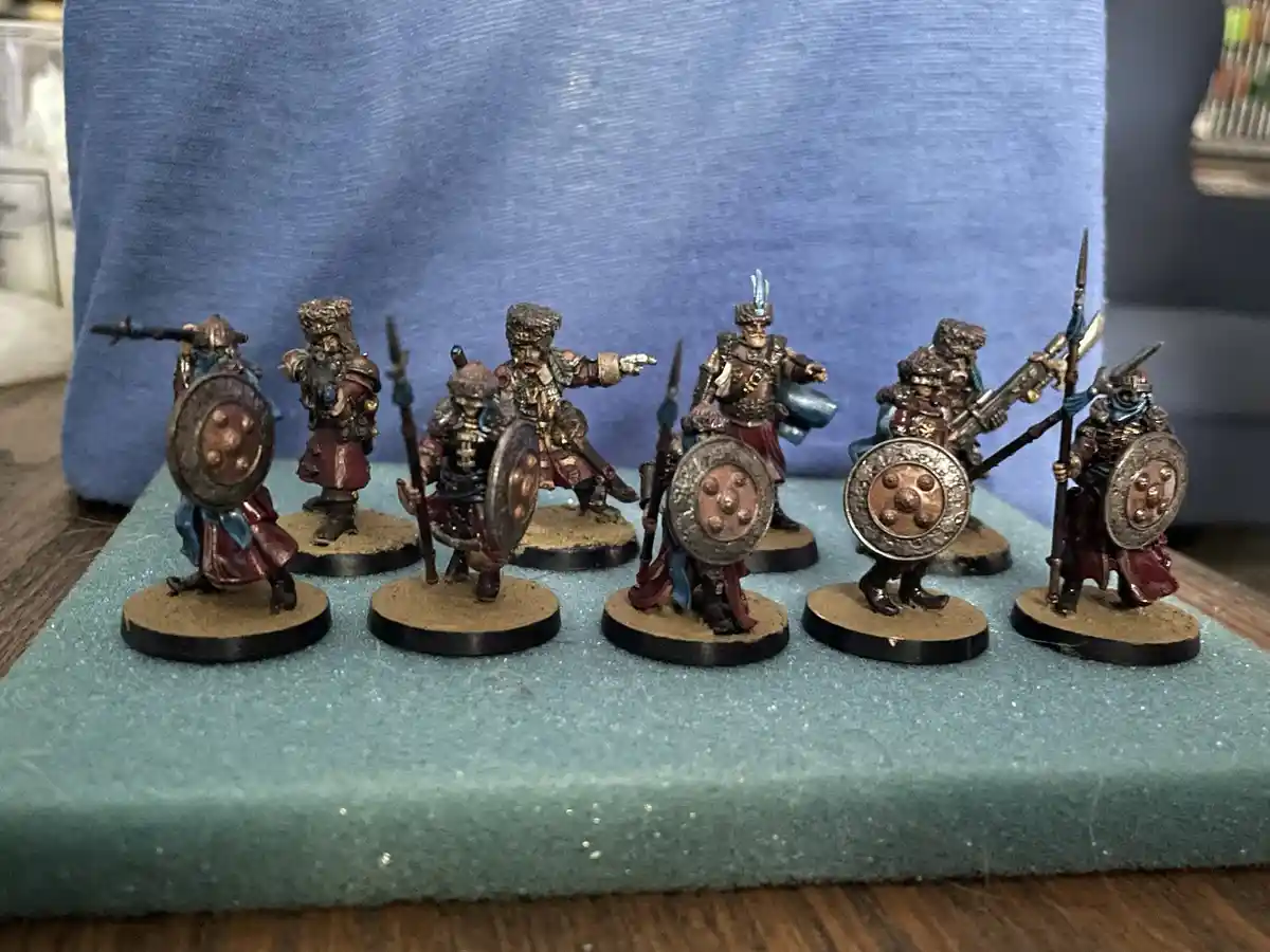 A row of seven hand-painted fantasy miniatures, resembling ancient warriors or barbarians with shields, spears, and armor…