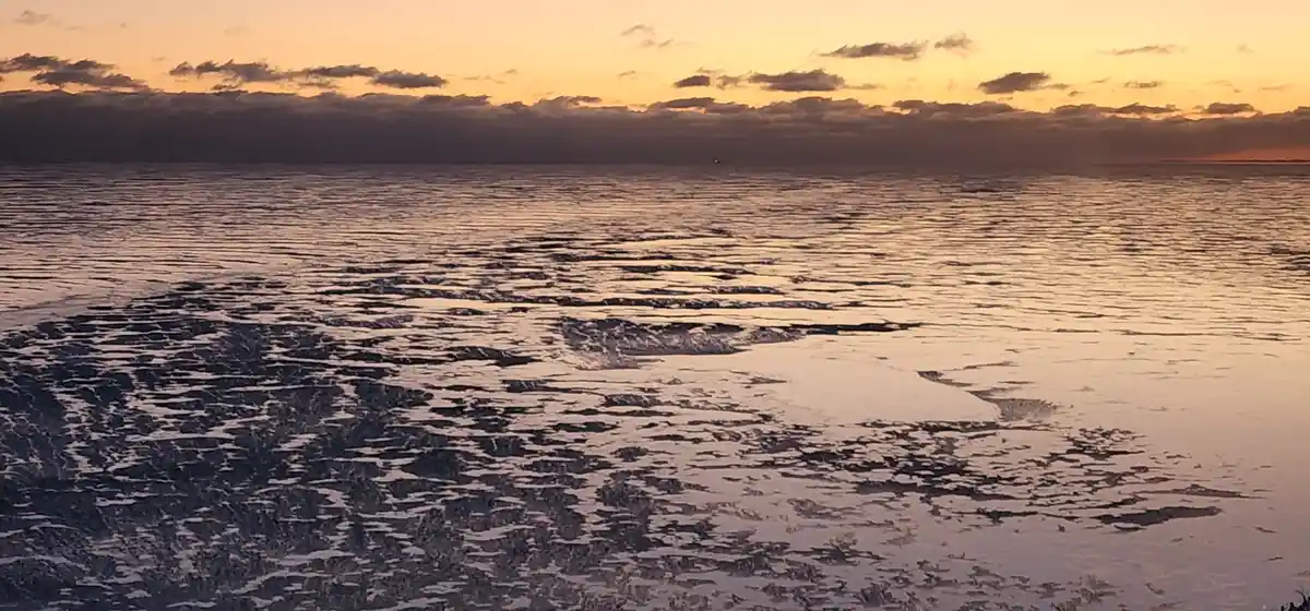 A serene sunset over a calm ocean with scattered ice floes in the foreground, reflecting the orange and yellow sky. The…