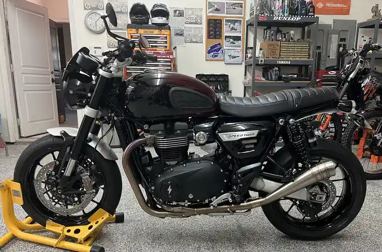 A black and maroon Triumph Speed Twin motorcycle is parked indoors on a yellow stand, with a British flag emblem on its fuel…