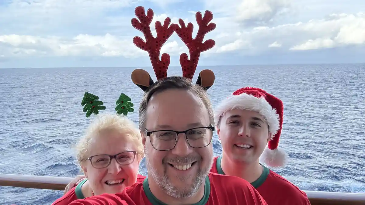 Three people on a cruise ship deck, wearing festive holiday headgear including reindeer antlers and a Santa hat, smiling for…