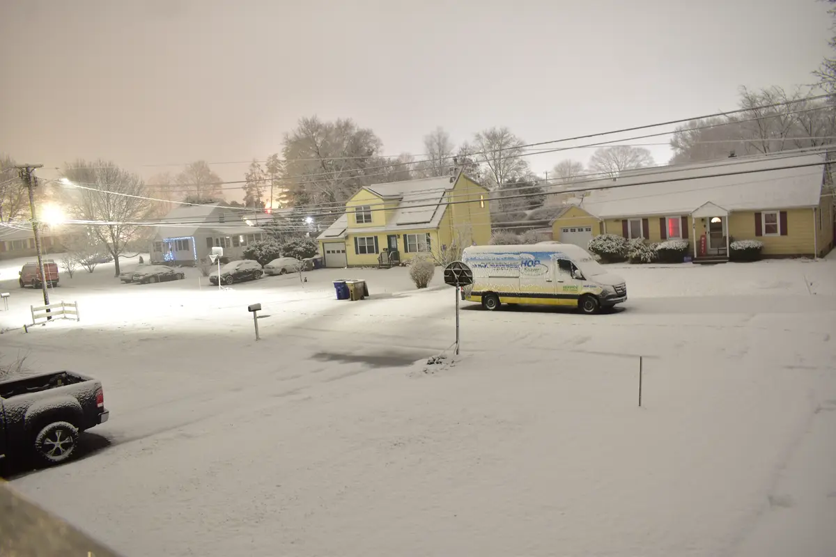 A snowy suburban street at night with houses, parked cars, and a snow-covered van with 'HOP Electric' branding. The van is…
