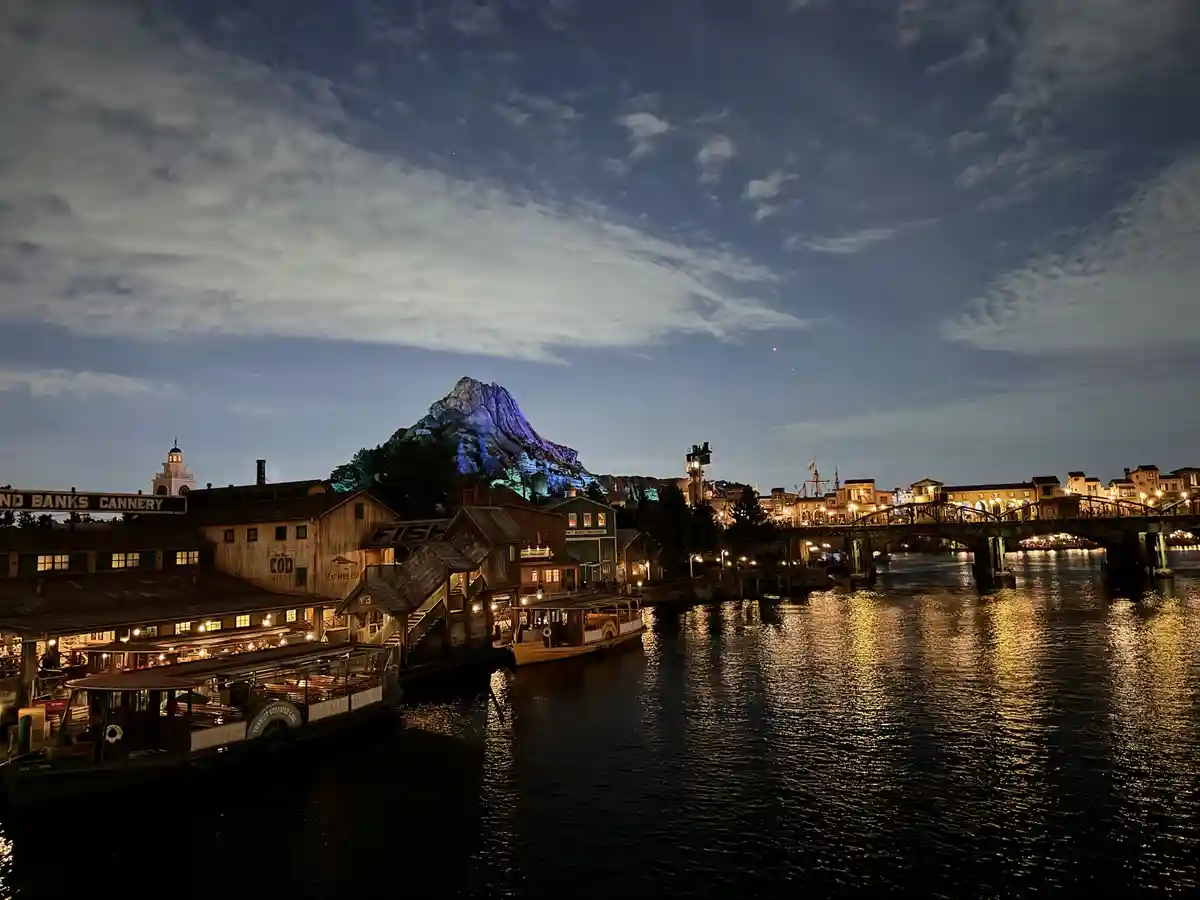A nighttime photo of a waterfront town with wooden buildings, boats, and a large, illuminated mountain in the background…