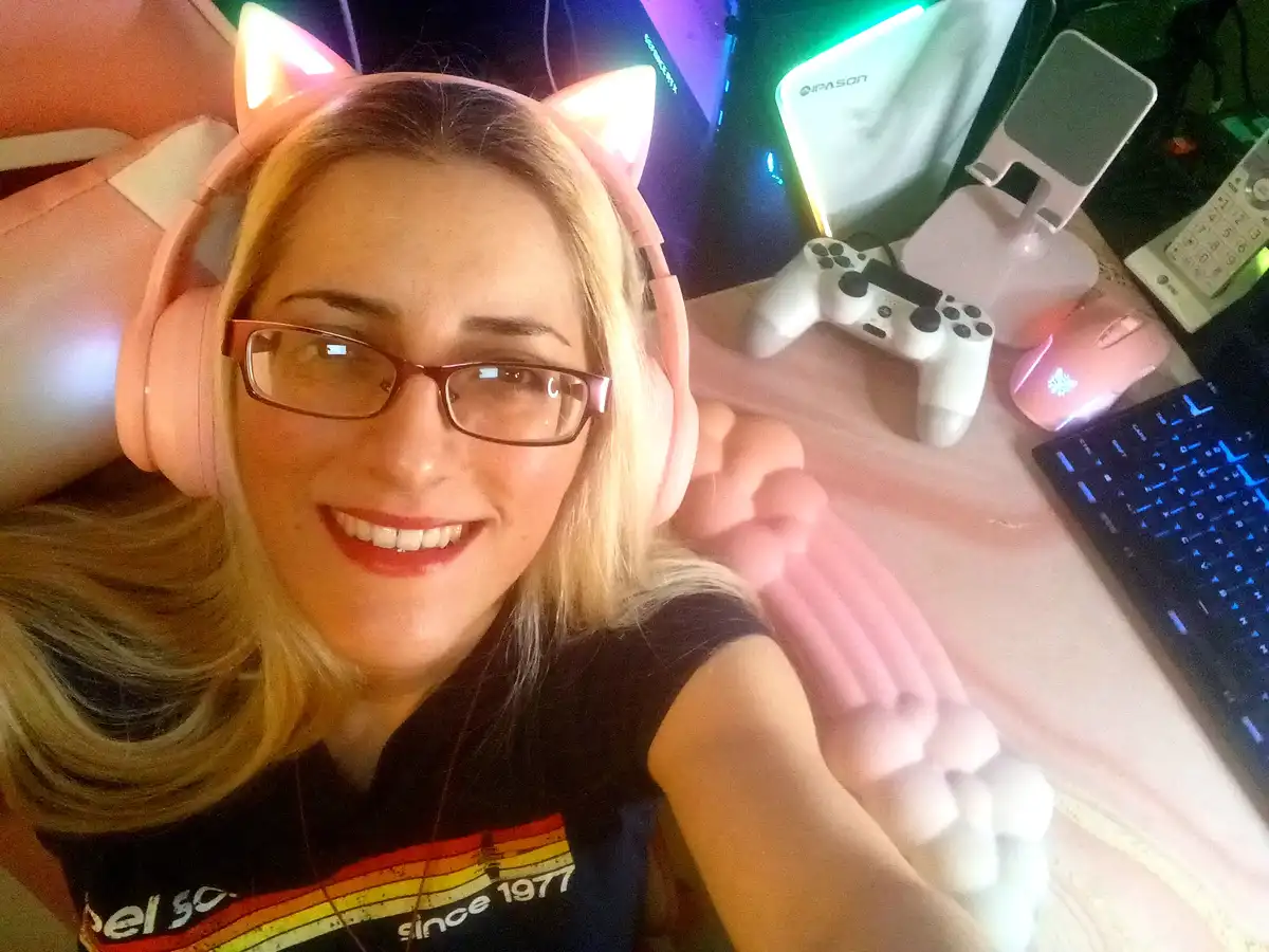 A smiling woman with blonde hair and glasses wears pink cat-ear headphones and waves at the camera. Behind her is a gaming…