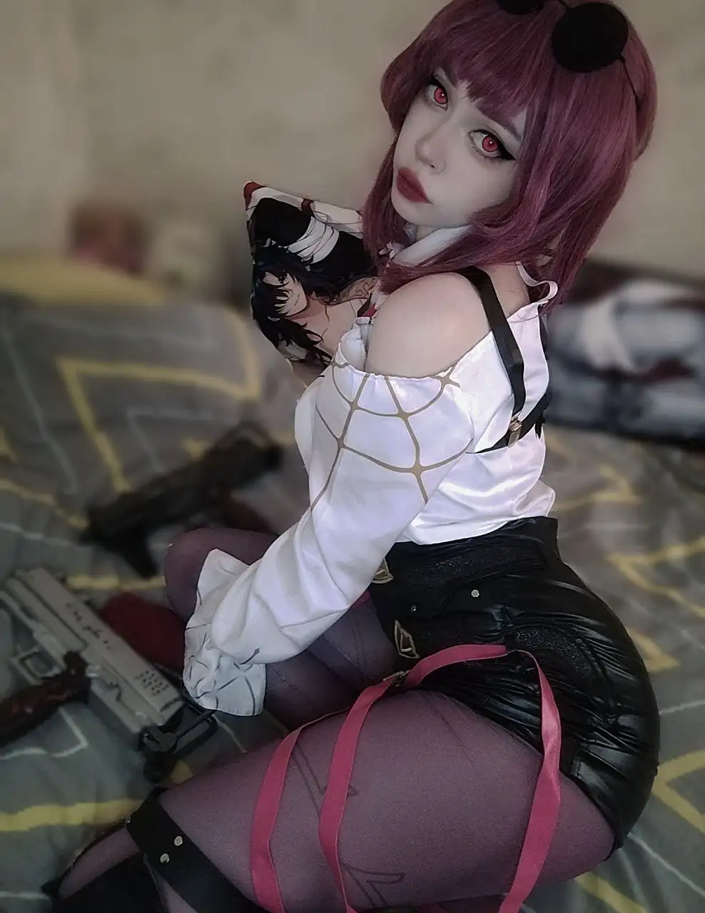 A person in cosplay with purple hair, red eyes, and a white top with black straps sits on a bed holding a pillow with an…