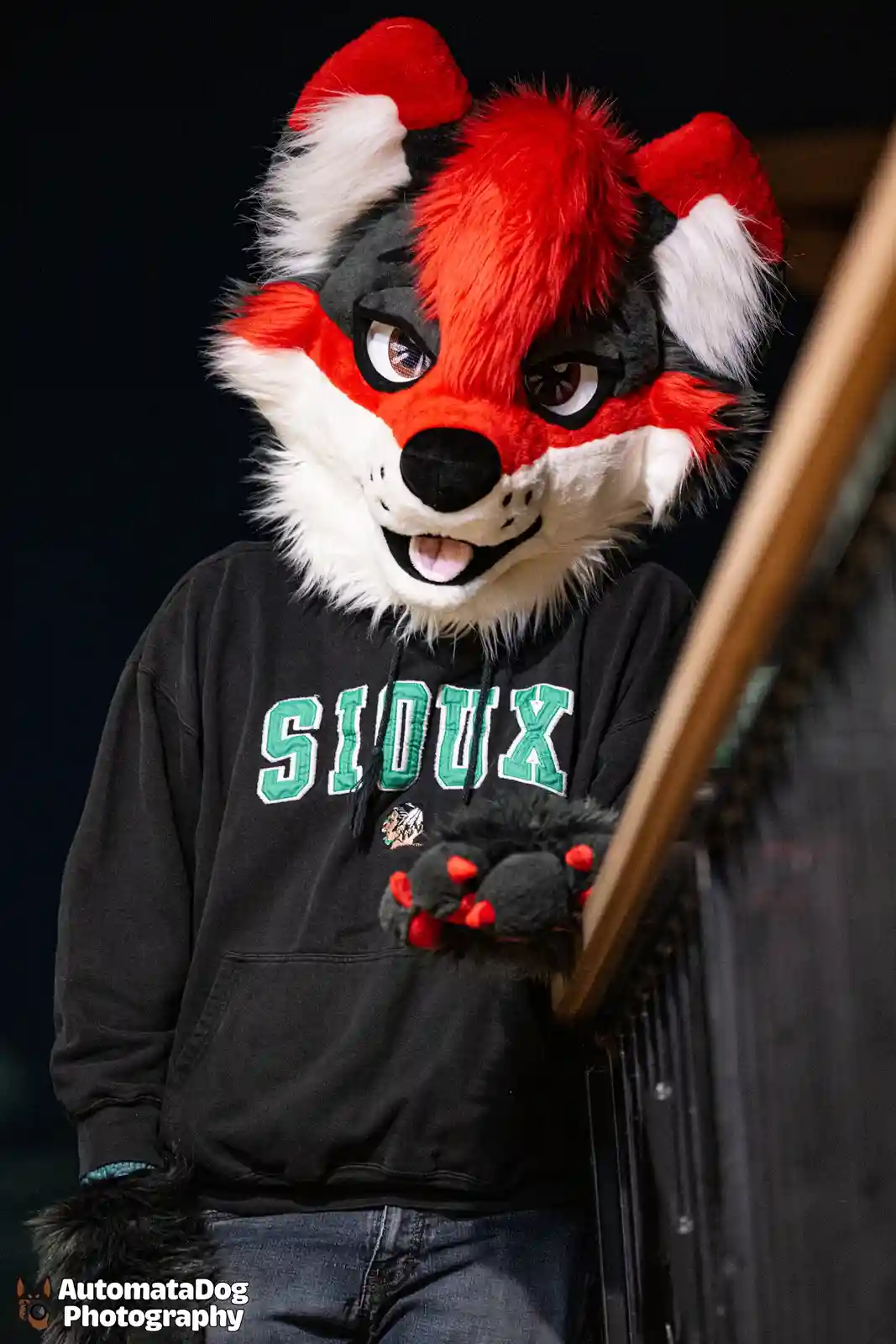 A person in a red, black, and white fox fursuit wearing a black hoodie with 'SIoux' in green letters, leans against a…