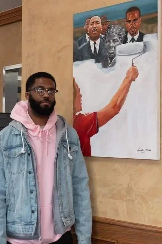 A man in a pink hoodie and denim jacket stands beside a large painting depicting a person painting over the faces of…