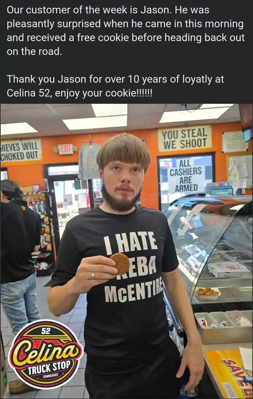 A man with a beard and brown hair stands inside a truck stop, holding a small cookie. He wears a black t-shirt with white…
