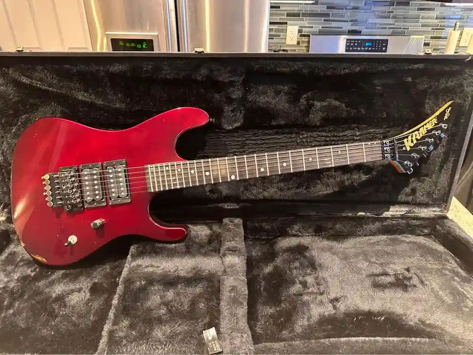 A red Kramer electric guitar rests inside its open black plush case, positioned in a kitchen with stainless steel appliances…