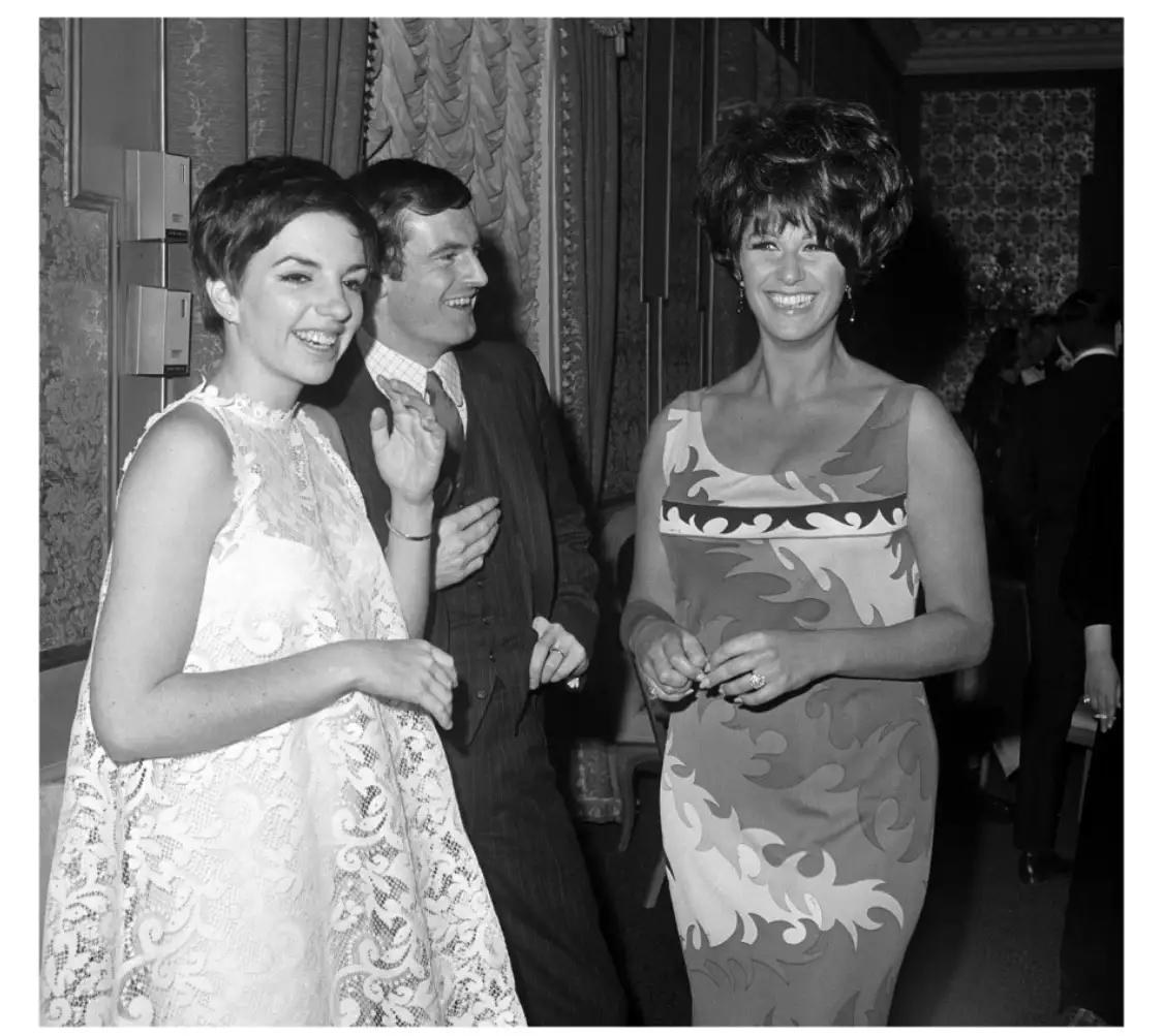 A black-and-white photo from the 1960s shows three people at a social gathering: a woman in a lace dress on the left, a man…