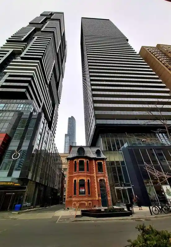 A low-angle photo shows a tiny, ornate red-brick Victorian house standing between two massive, modern glass-and-steel…