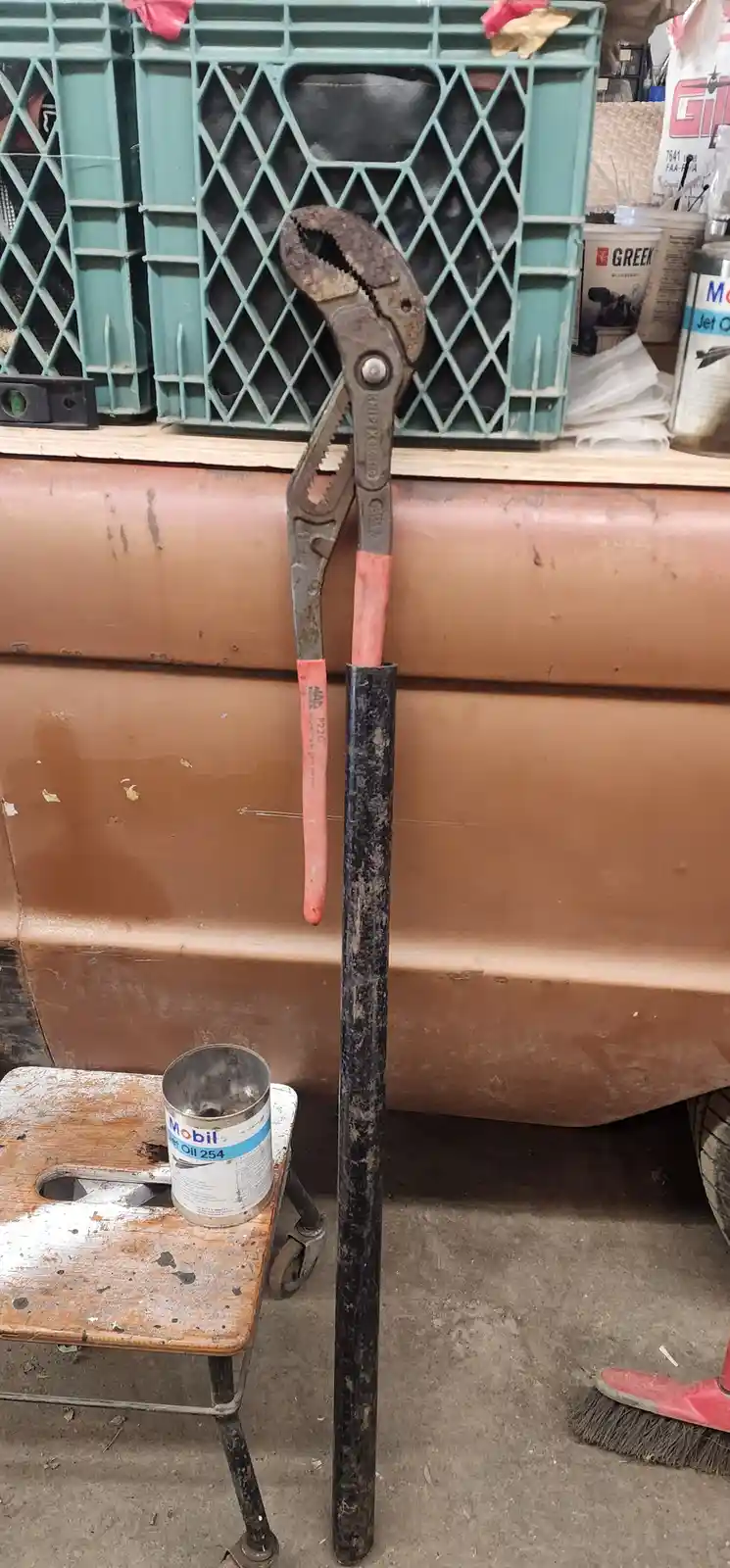A large, rusty pair of channel lock pliers with red handles is stuck in a black metal pipe, standing upright in a cluttered…