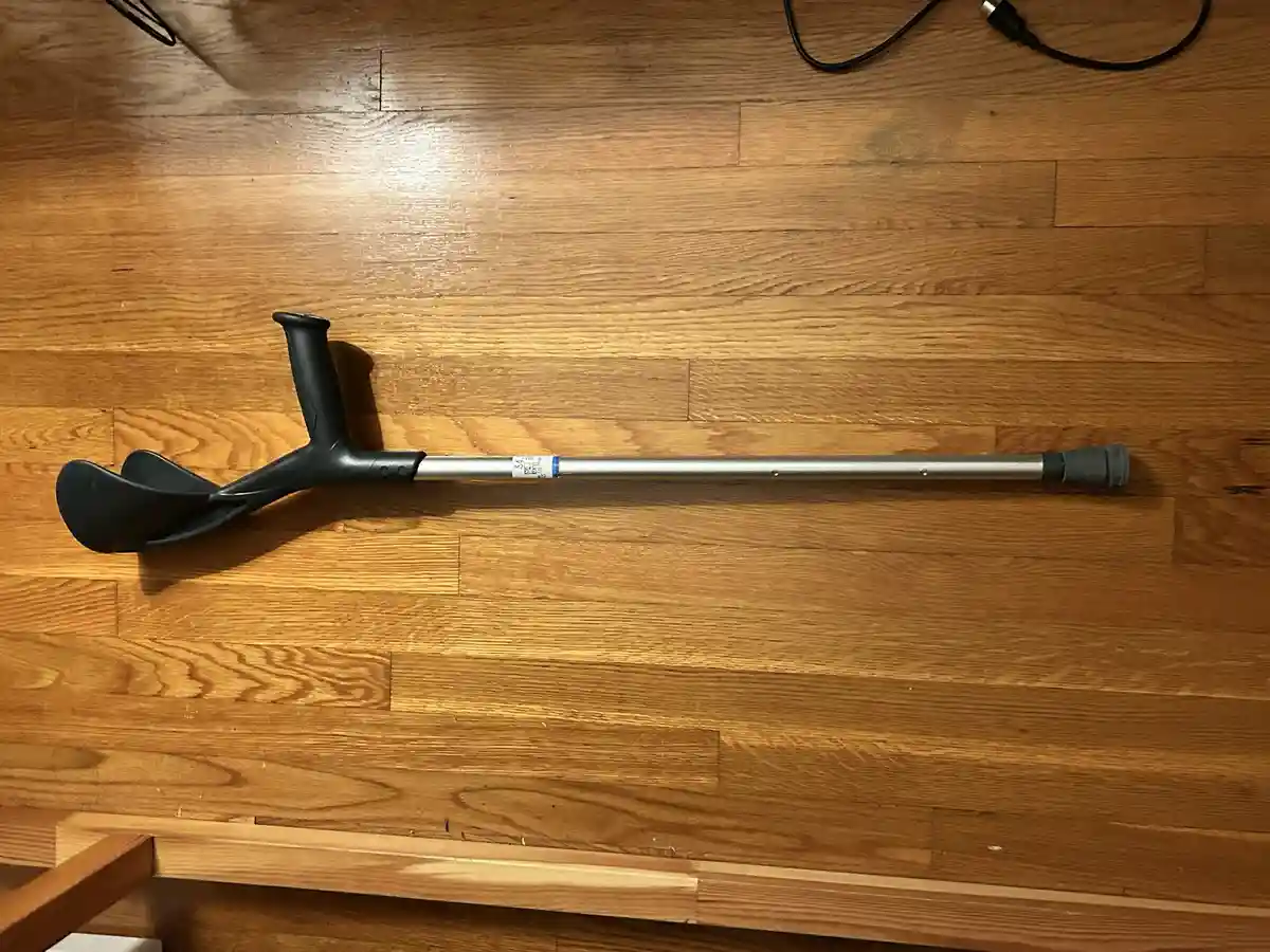 A black and silver adjustable crutch lies horizontally on a light brown wooden floor, with its padded handgrip and rubber…