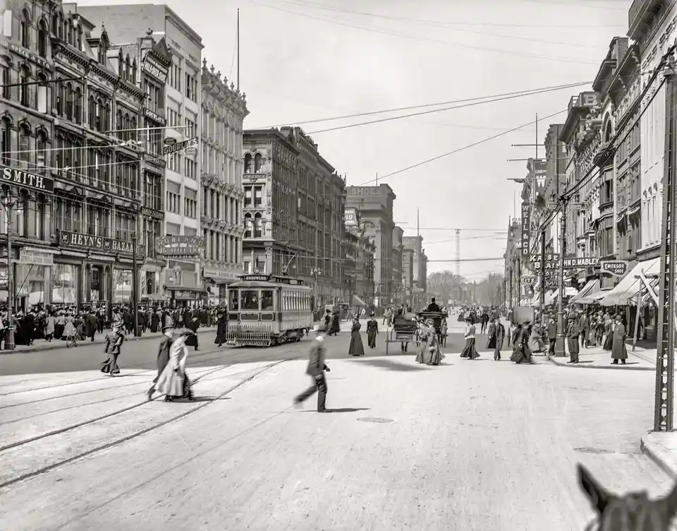 A vintage black-and-white photograph shows a busy, crowded city street from the early 1900s, with pedestrians, horse-drawn…