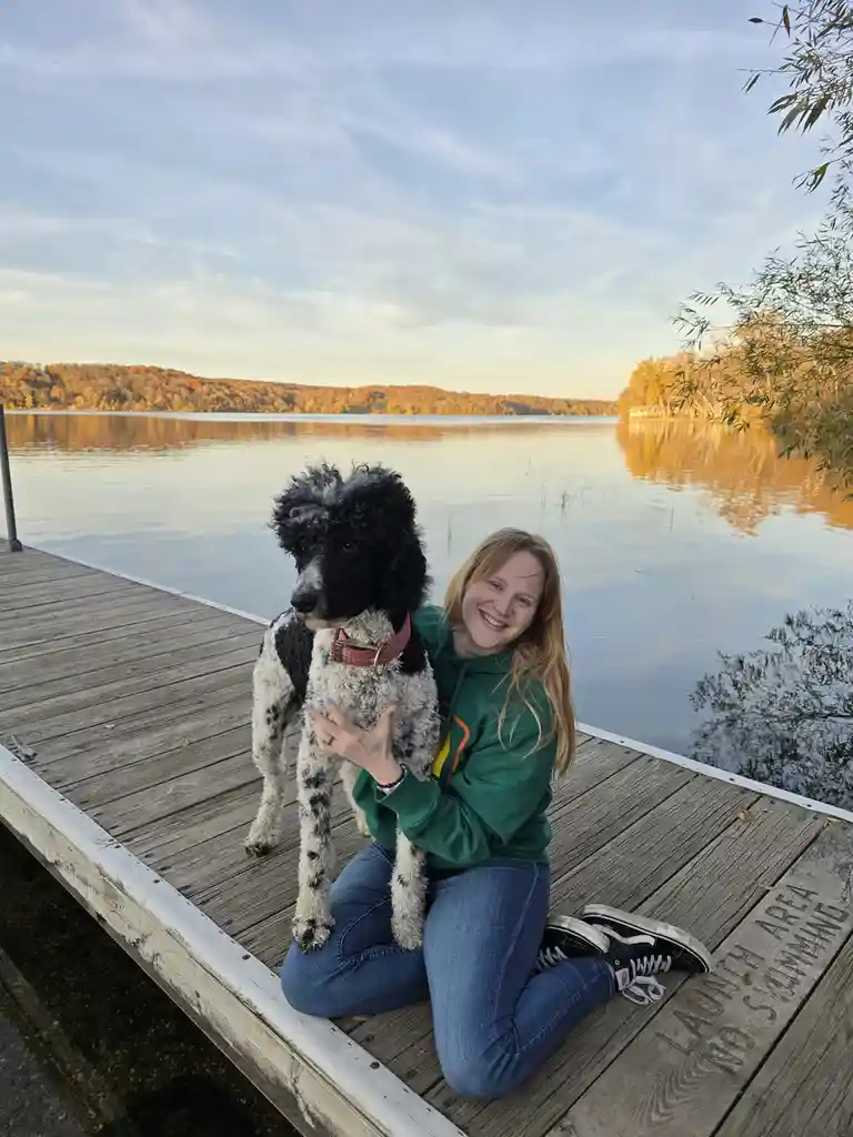 A smiling woman in a green hoodie kneels on a wooden dock next to a black-and-white curly-haired dog, both looking at the…