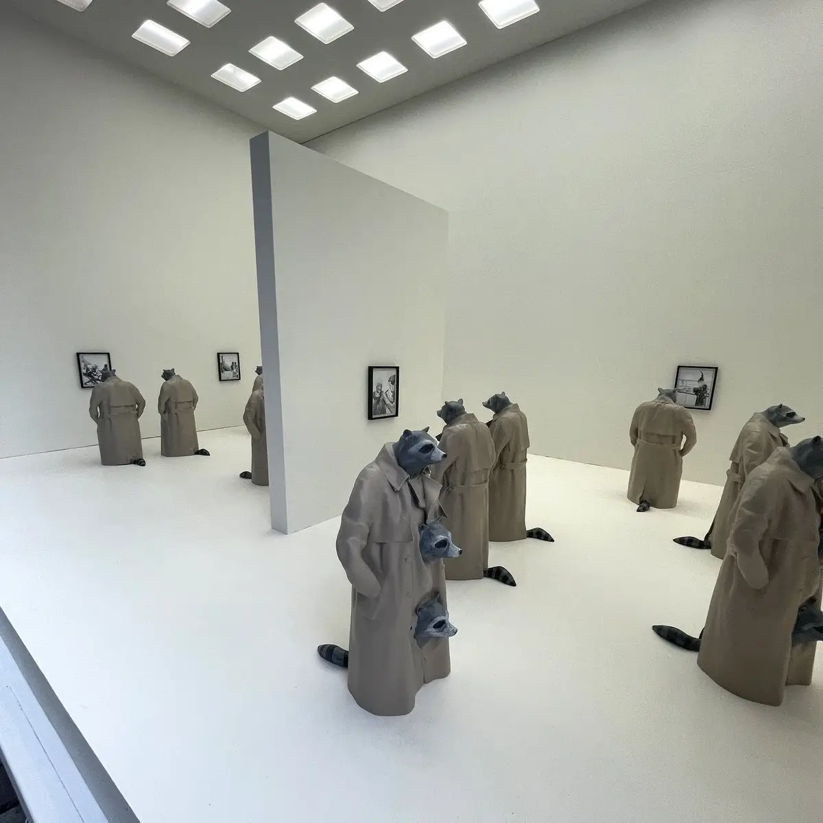 The image shows multiple sculptures of raccoons wearing beige trench coats, standing in a minimalist white gallery space and…