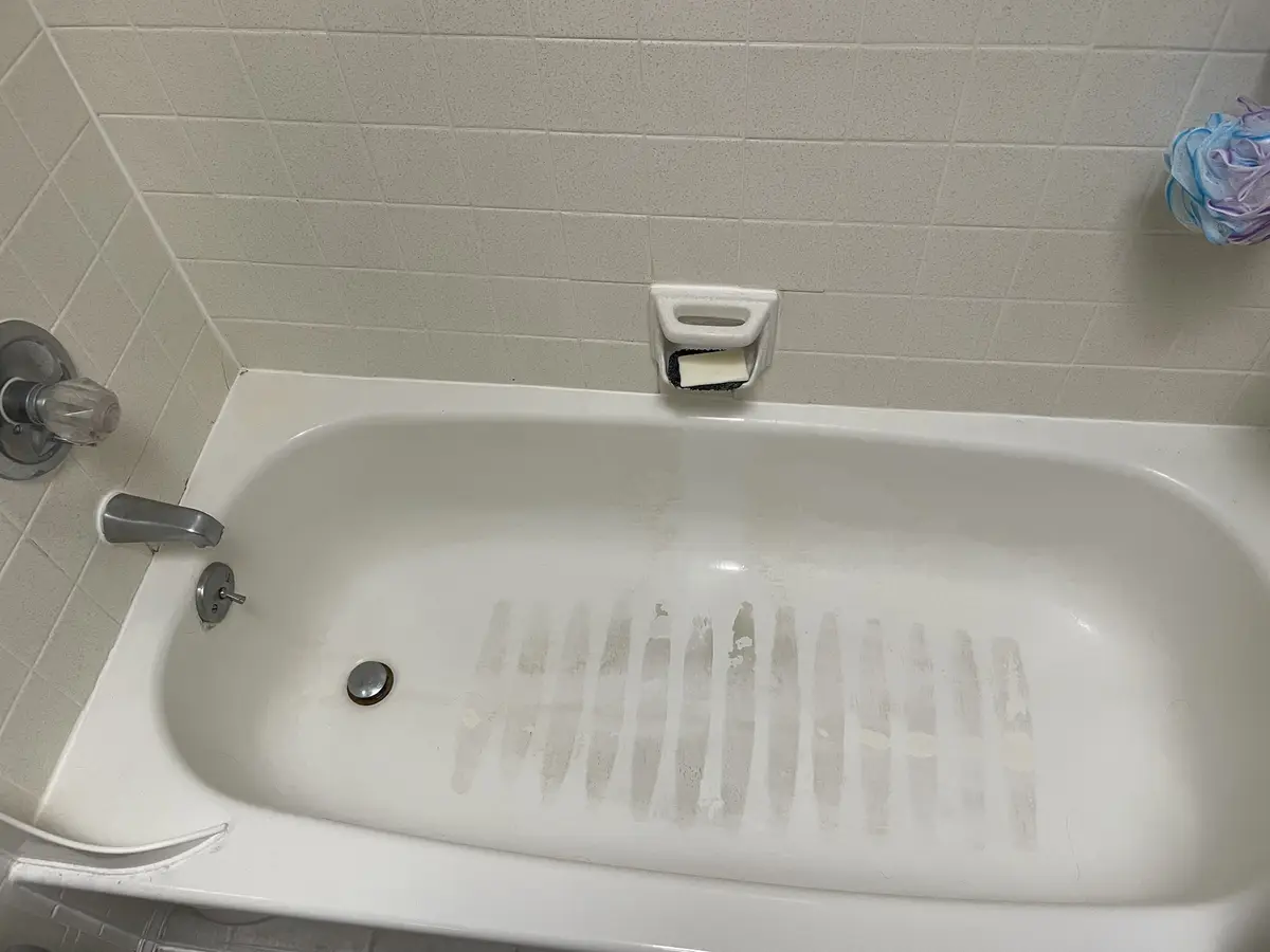 A white bathtub with light-colored tiled walls shows distinct, parallel streaks of cleaning residue across its bottom…
