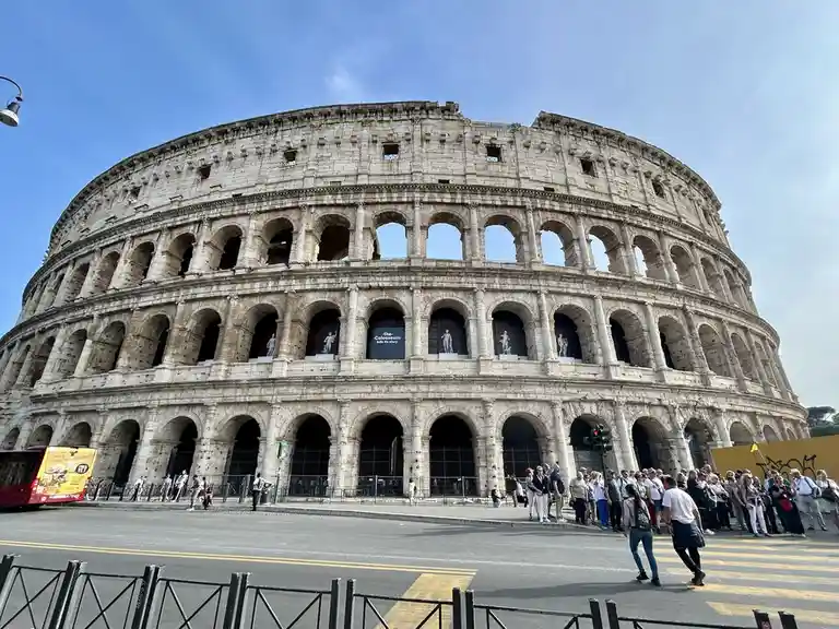 A wide-angle photo shows the massive, ancient Colosseum in Rome under a clear blue sky, with tourists gathered on the street…