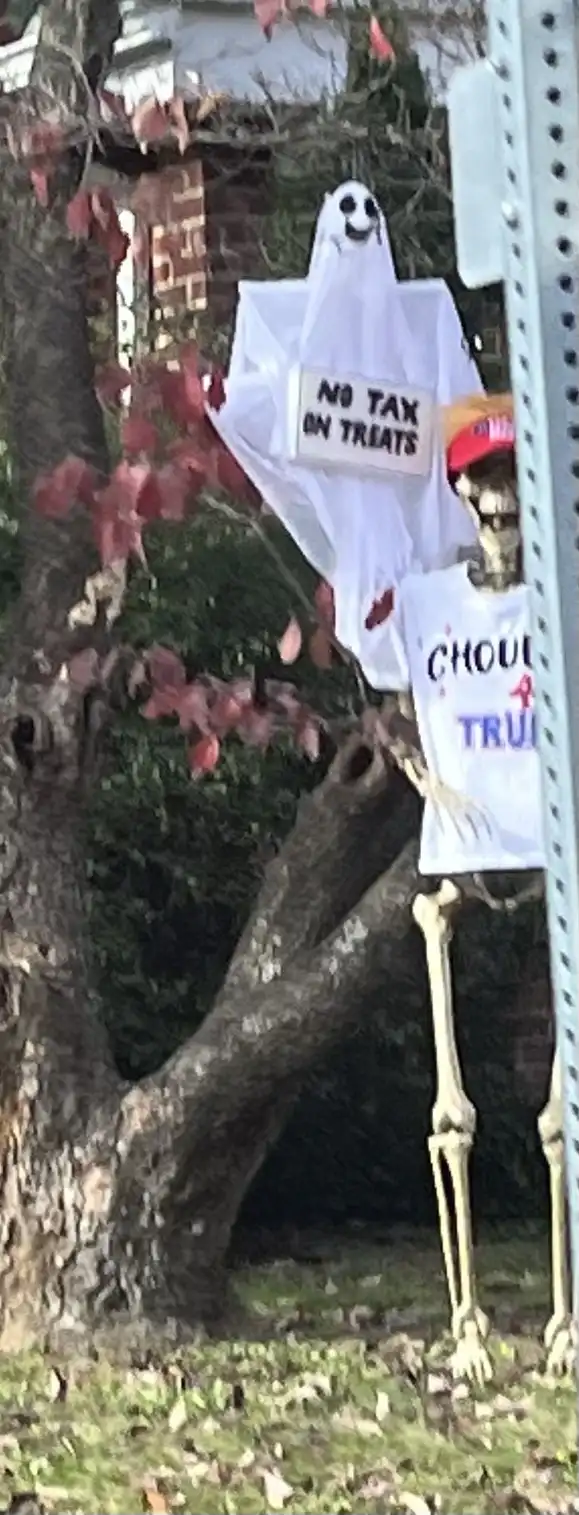 A Halloween decoration shows a ghost holding a sign that reads 'NO TAX ON TREATS' while a skeleton wearing a red MAGA hat…