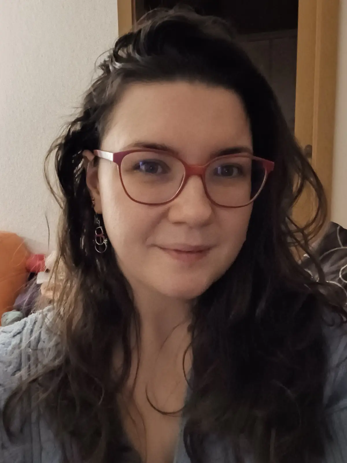 A close-up selfie of a woman with dark, wavy hair, wearing red-framed glasses and dangling earrings, smiling slightly while…