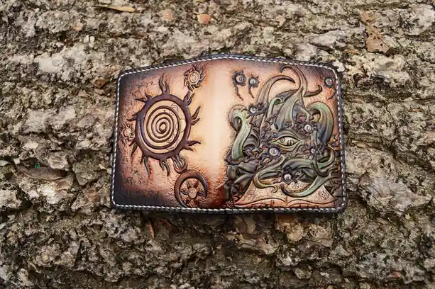 A handcrafted leather wallet with intricate embossed designs rests on rough tree bark. The left side features stylized suns…