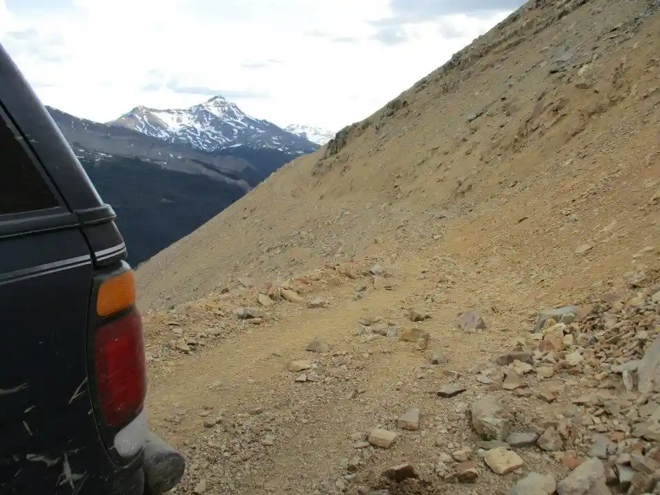 A black SUV is parked on a steep, rocky, dirt mountain trail with snow-capped peaks visible in the distance. The overlaid…