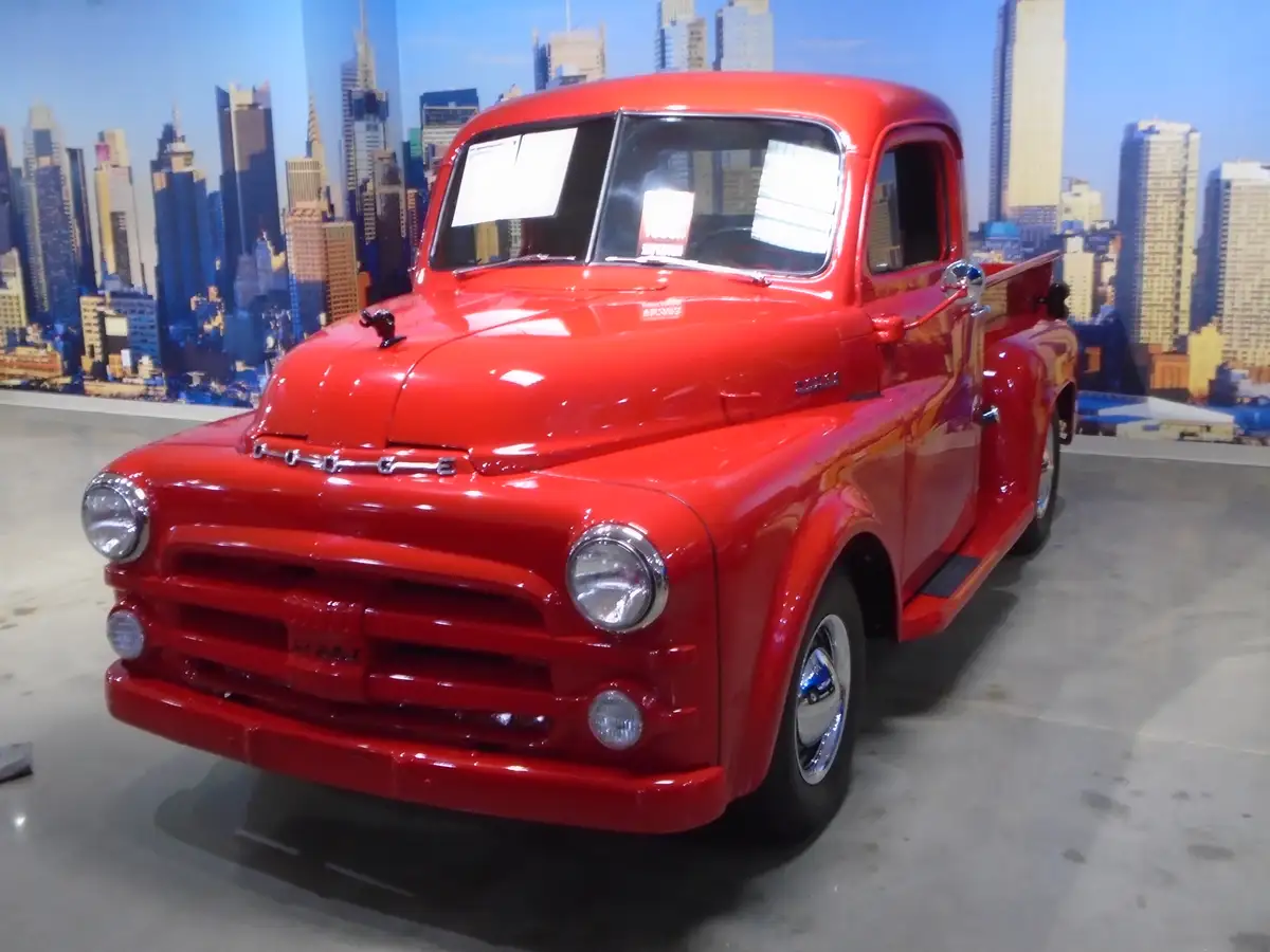 A shiny red vintage Dodge pickup truck is displayed indoors in front of a large city skyline backdrop. The truck has chrome…