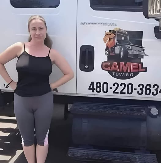 A woman in a black tank top and grey leggings stands confidently with hands on hips beside a white tow truck. The truck's…